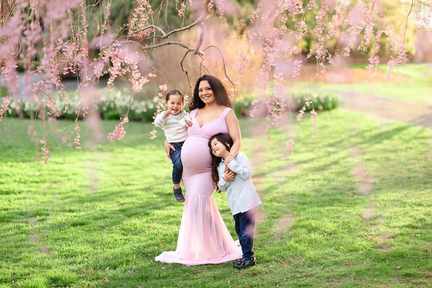 Two siblings cuddle with their mother beneath cascading cherry blossoms during a maternity session in Bristol, Rhode Island.