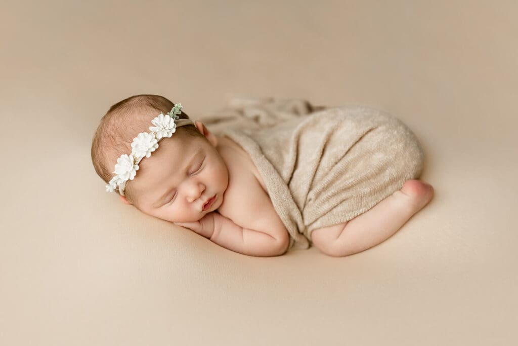 Sleeping newborn baby wrapped in a neutral toned fabric with a floral headband during an in home newborn photography session in Cumberland, Rhode Island