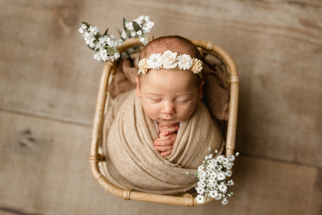 Wrapped newborn baby sleeping in a bamboo basket with white florals and a floral headband during an in home newborn photography session in Cumberland, Rhode Island