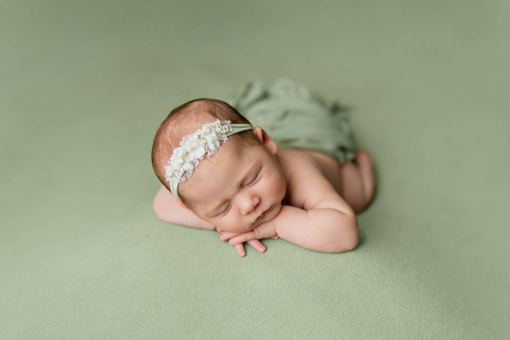 Sleeping newborn girl in chin on hands pose on sage green backdrop with floral headband during posed newborn session in Cumberland Rhode Island