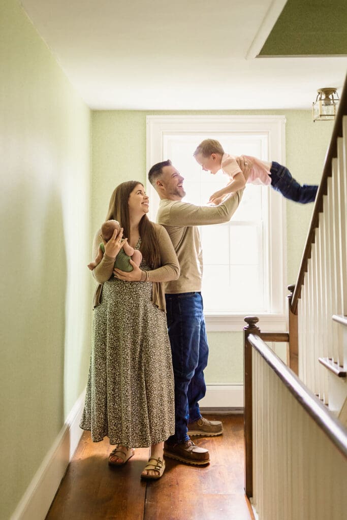 Father playfully swinging his son on the staircase while mother holds their newborn during an in home lifestyle newborn photography session in Cumberland, Rhode Island
