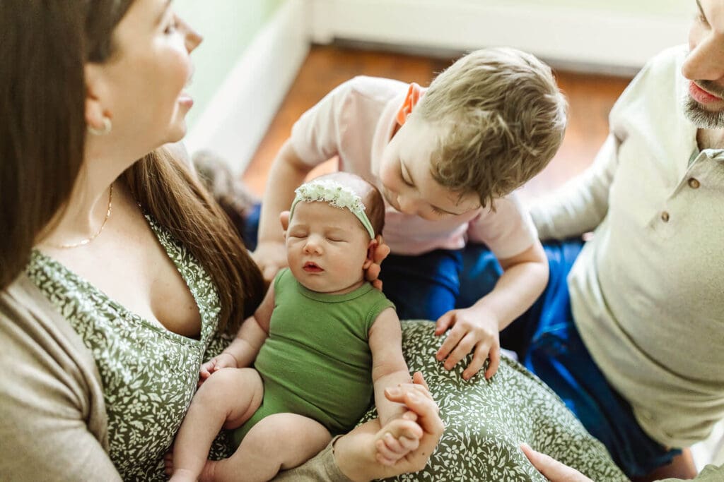 Older sibling kissing his newborn sister on the cheek while mom cradles her during an in home lifestyle newborn photography session in Cumberland, Rhode Island
