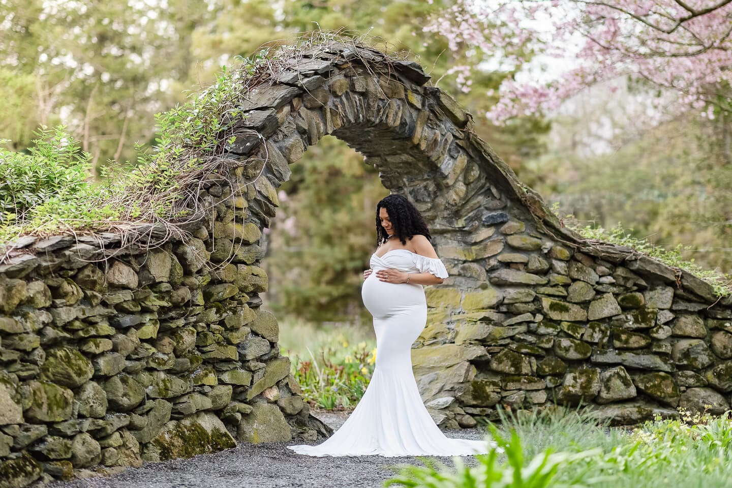 Expecting mother in a white maternity gown standing beneath a stone arch during a maternity photography session in Bristol, Rhode Island.