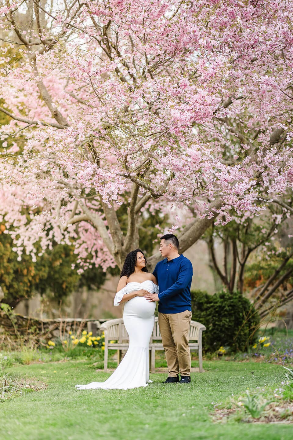Mother and father to be posing under pink flowering cherry trees during a maternity session in Bristol, Rhode Island.