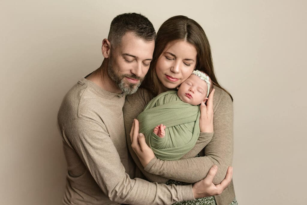 Parents embracing their newborn daughter against a neutral backdrop during an in home newborn photography session in Cumberland, Rhode Island