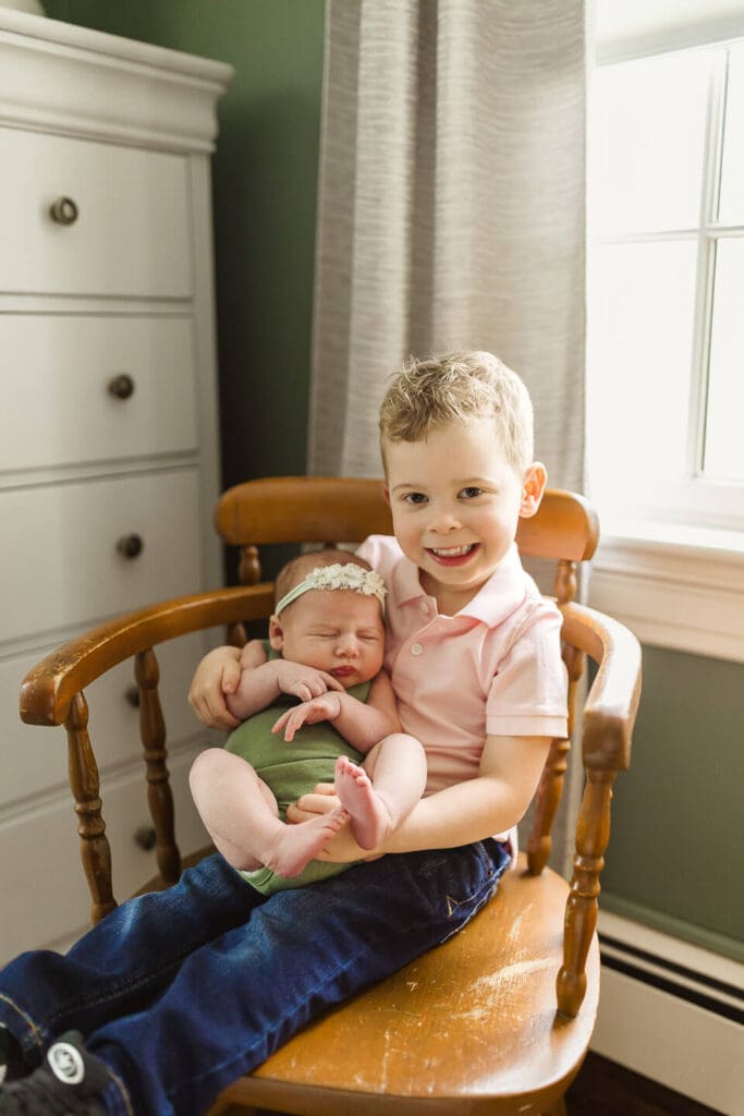 Young boy holding newborn sister in his bedroom during lifestyle newborn session in Cumberland Rhode Island