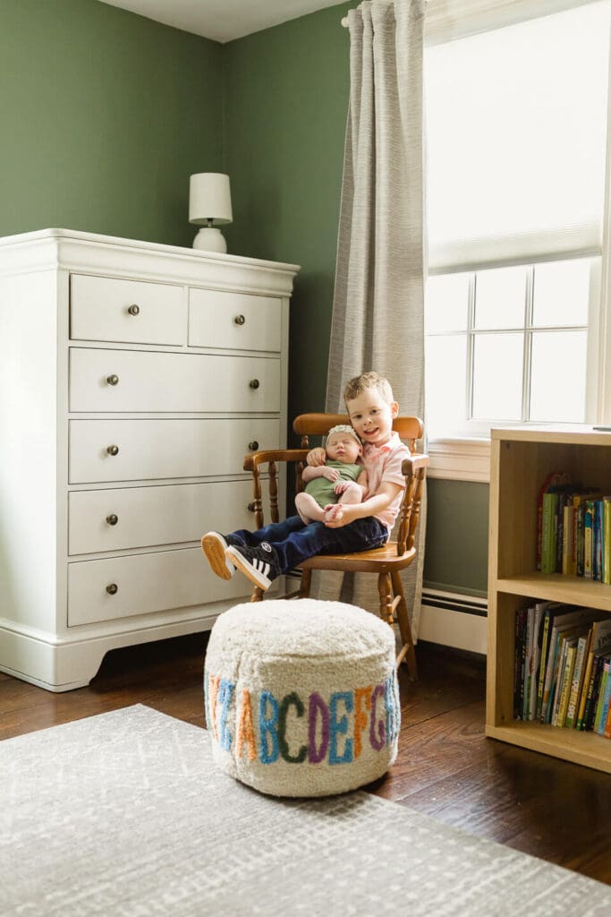 Young boy holding newborn sister in wooden chair in sage green nursery during lifestyle newborn session in Cumberland Rhode Island